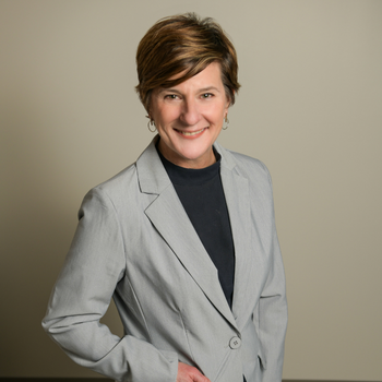 A white woman with cropped brown hair wearing a black top and a gray blazer