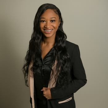 A black woman with long curly hair wearing a black top and a black and tan blazer.
